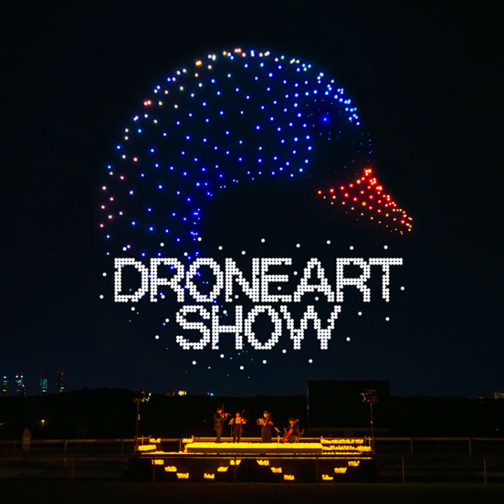 A night scene featuring a colorful drone light display forming the shape of a duck, with the text 'DRONE ART SHOW' illuminated below, accompanied by performers playing instruments.