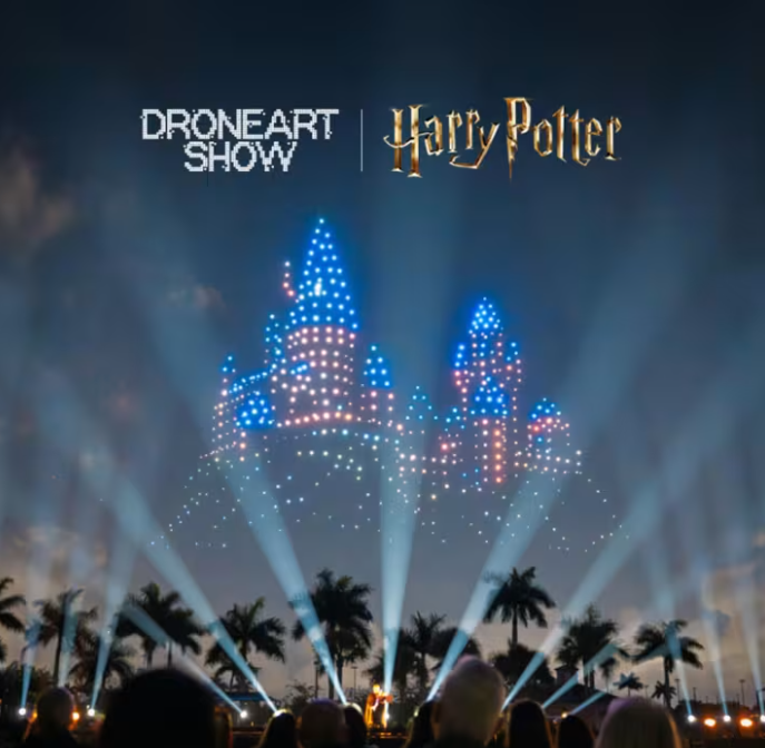 A drone light show featuring a castle illuminated with blue and pink lights, set against a twilight sky, with spectators in the foreground and palm trees around.